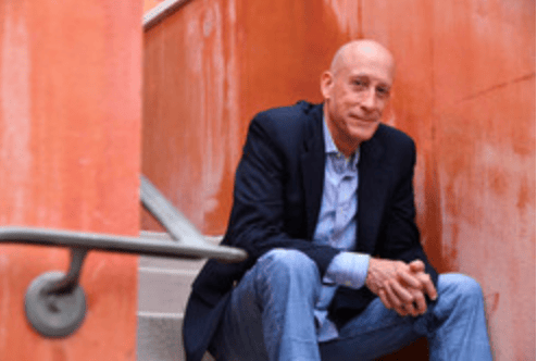 Matt Fraser, a bald man in a navy blazer and jeans, sits on stairs against an orange wall, looking at the camera with a slight smile.